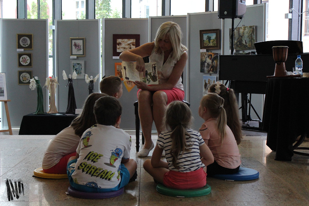 W holu Biblioteki Głównej siedzi na krześle prowadząca zajęcia – Wanda Kołodziejska. Czyta książkę wskazując palcem na obrazki przedstawiające instrumenty muzyczne. Na podłodze w półkolu siedzą dzieci.