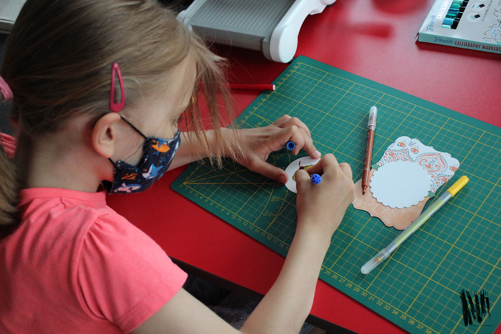 Przy stole siedzi dziewczynka i koloruje pisakami element przygotowywanej kartki z motywem babeczki.