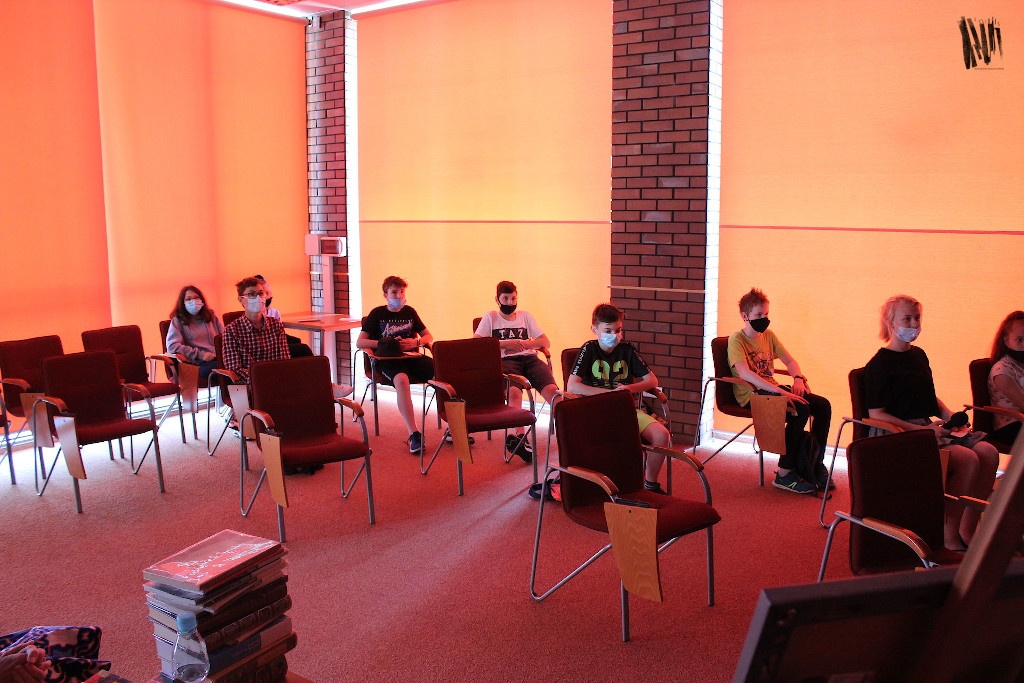 W sali o ciepłej, pomarańczowo-brązowej kolorystyce siedzą na krzesłach uczniowie biorący udział w spotkaniu.