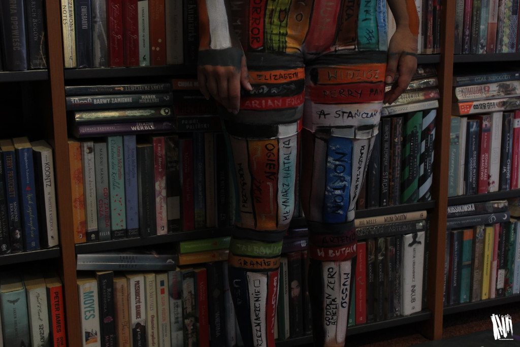 Przy regale z książkami stoi postać, widziana od pasa w dół, całkowicie wtapiająca się w tło, przez pomalowane na ciele kolorowe paski, imitujące okładki książek.