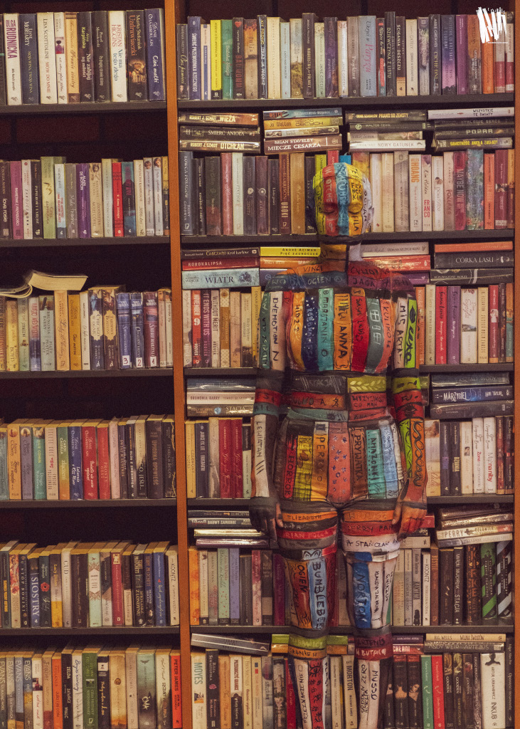Przy regale z książkami stoi modelka całkowicie wtapiająca się w tło, przez pomalowane na ciele kolorowe paski, imitujące okładki książek.