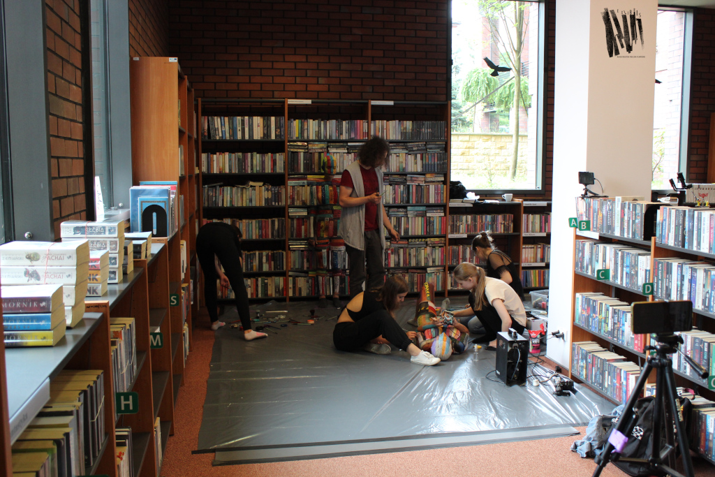 W Bibliotece Głównej, przy narożnych regałach, grupa osób maluje leżącą na podłodze modelkę. Druga modelka, odpowiednio pomalowana, stoi przy regale z książkami niemal całkowicie komponując się z tłem.