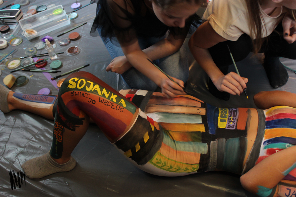 Na folii rozłożonej na podłodze leży modelka, której ciało malują dwie osoby w kolorowe pasy. Obok znajdują się pojemniki z farbami.