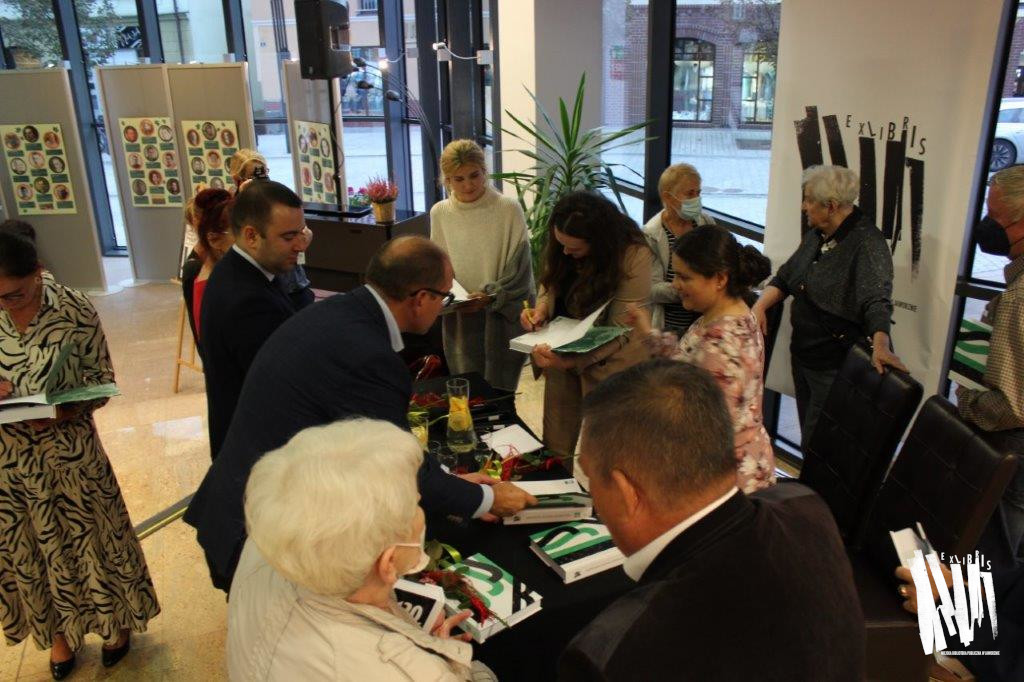 Hol Biblioteki, przy stoliku stoją i rozdają autografy autorzy Jaworznickiego Słownika Biograficznego wokół zebrali się uczestnicy spotkania.