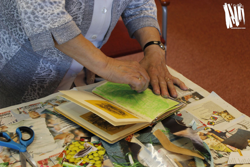 Zbliżenie na dłonie uczestnika zajęć w trakcie pracy nad ozdabianiem książki, wokół przygotowane do przyklejenia wycinki z gazet. 