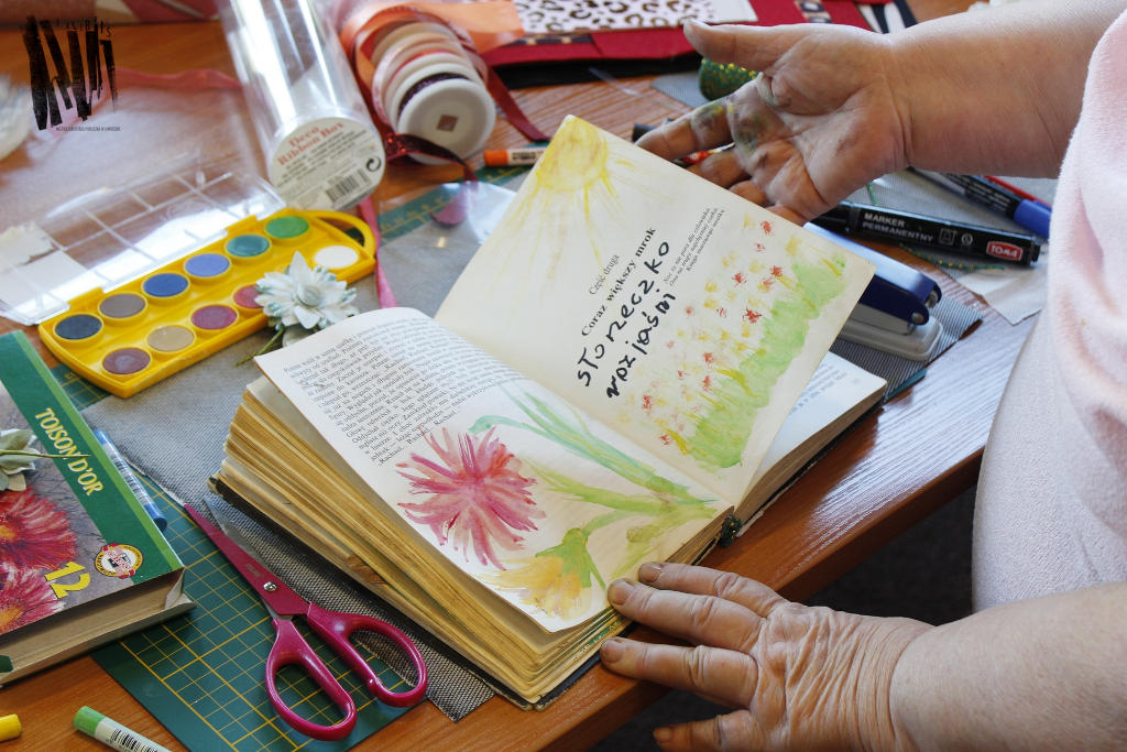 Na stole w dłoniach otwarta książka z namalowanymi kwiatami, wokół materiały plastyczne i artystyczne: farby, nożyczki, markery, wstążki. 