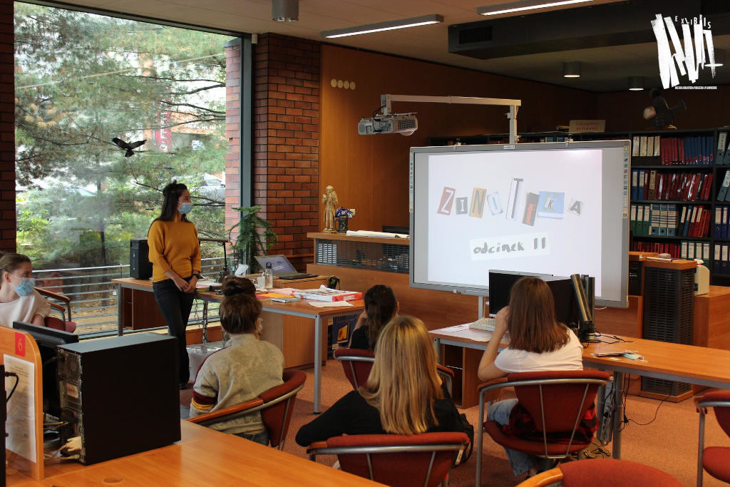 Biblioteka Główna w Jaworznie, oszklona przestrzeń Czytelni Naukowej, uczestnicy warsztatów wraz z prowadzącą patrzą na prezentację wyświetlaną na tablicy interaktywnej.