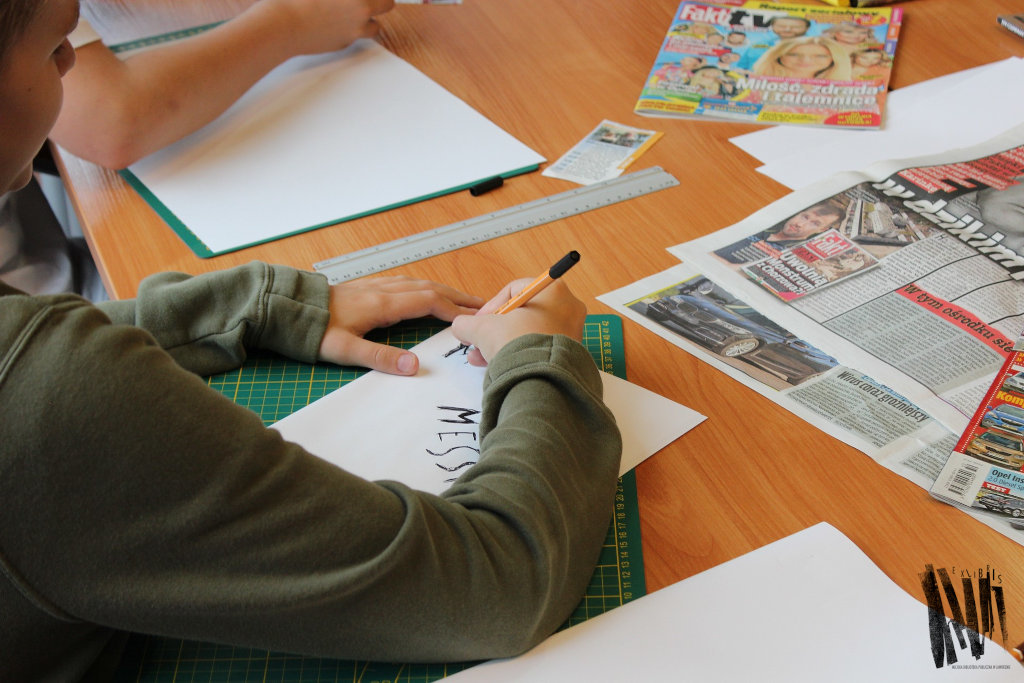 Uczestnik warsztatów podczas pracy: zbliżenie na dłonie, rysowanie cienkopisem, wokół rozłożone na stole gazety i materiały plastyczne.