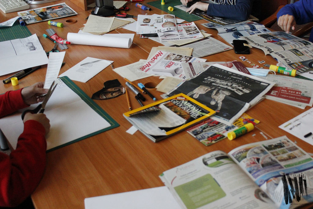 Uczestnicy warsztatów podczas pracy: wycinanie elementów z gazet, wokół rozłożone na stole czasopisma i materiały plastyczne.