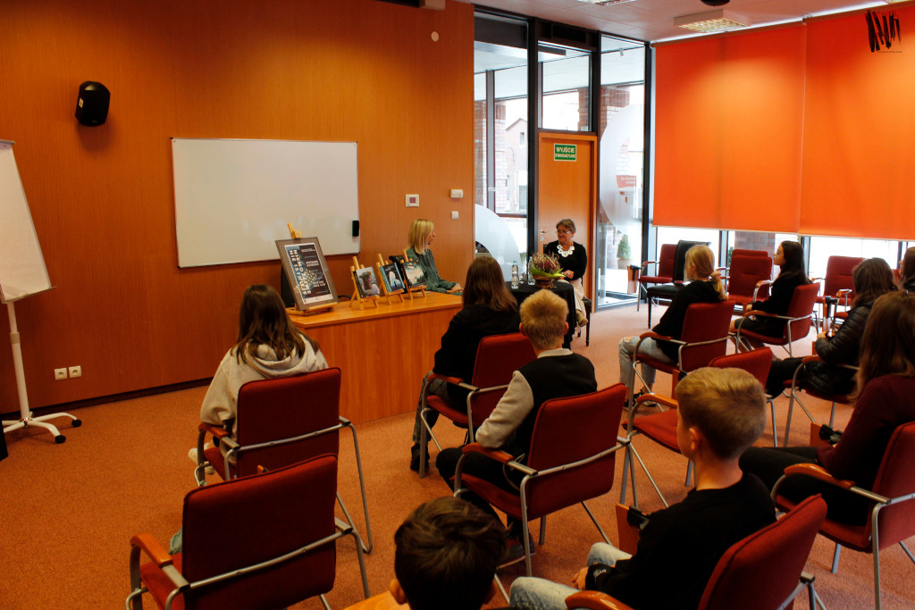 Na kolorowym zdjęciu widzimy siedzącą pisarkę Agnieszkę Kaźmierczyk, która rozmawia z prowadzącą spotkanie kobietą. Paniom przysłuchuje się młodzież zaproszona na spotkanie.