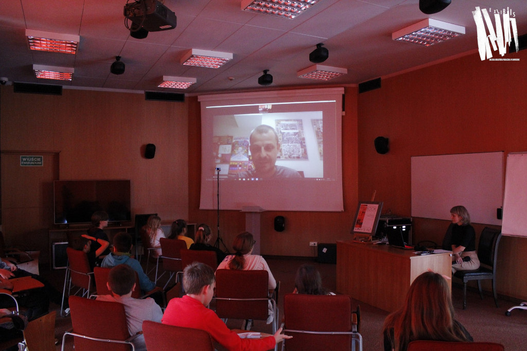 Audytorium Biblioteki Głównej w Jaworznie, na dużym ekranie twarz Łukasza Kowalczuka, na pierwszym planie siedzący na krzesłach uczestnicy spotkania wraz z pracownikiem biblioteki.