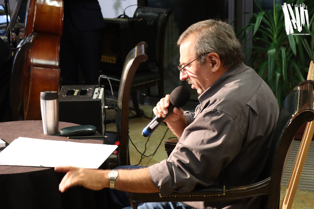 Dominik Socha, starszy mężczyzna w okularach, ubrany w szarą koszulę, siedzi przy niewielkim stoliku. Mężczyzna pochyla się nad zapisaną kartką leżącą przed nim na okrągłym stoliku i mówi do trzymanego w prawej dłoni mikrofonu.
