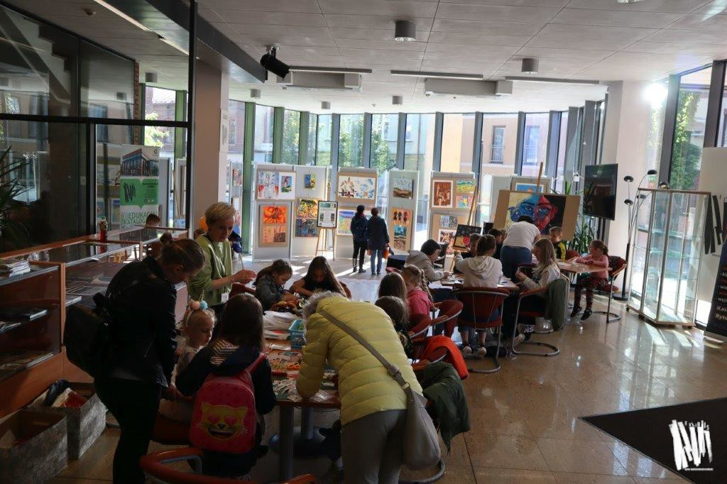 W holu Biblioteki Głównej, niedaleko Galerii ExLibris, ustawiono dwa duże stoły. Przy każdym stole siedzi grupa dzieci, a za ich plecami stoją rodzice i opiekunowie.
