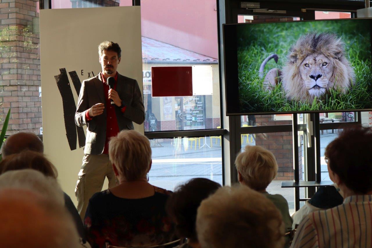 Na tle tłumnie zgromadzonej publiczności stoi Tomasz Michniewicz, który żywo gestykuluje, opowiadając o okolicznościach wykonania zdjęcia, które wyświetla się na ekranie. Fotografia przedstawia dużego lwa o płowej grzywie, który stoi wśród bujnych zielonych traw.