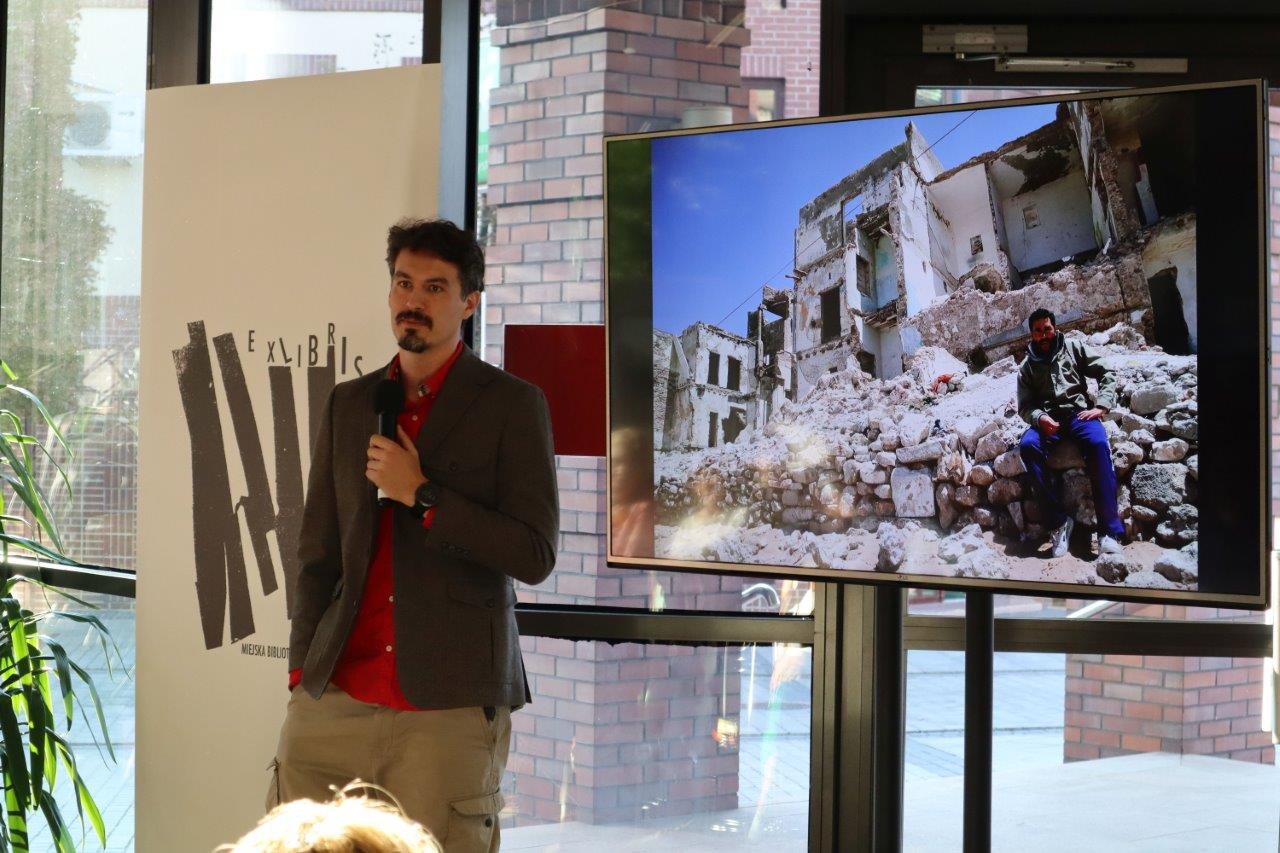 Na tle roll up’u z ekslibrisem MBP stoi Tomasz Michniewicz, który w jednej dłoni trzyma mikrofon, a drugą wsunął do kieszeni. Podróżnik spogląda wyczekująco na niewidocznych na zdjęciu widzów, którym przed momentem zadał pytanie na temat tego, z czym kojarzy im się fotografia wyświetlona na ekranie. Zdjęcie przedstawia zniszczony budynek i ciemnowłosego mężczyznę, który siedzi na rozrzuconym wszędzie gruzie.